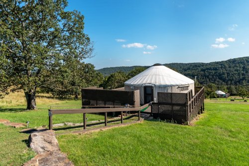 A yurt surrounded by green grass and trees, with a wooden ramp leading to its entrance against a hilly backdrop.