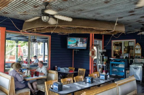 Interior of a casual restaurant with a canoe on the ceiling, tables set for dining, and a TV showing the weather.