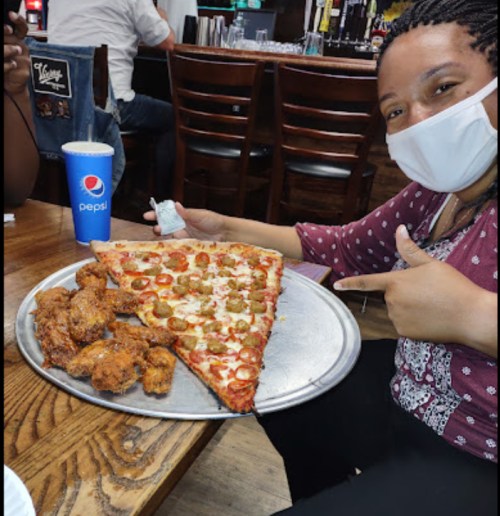 A person wearing a mask smiles and holds a large pizza slice and a plate of chicken wings at a restaurant.