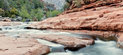A serene river flows over smooth, reddish rocks, surrounded by lush greenery and steep canyon walls.