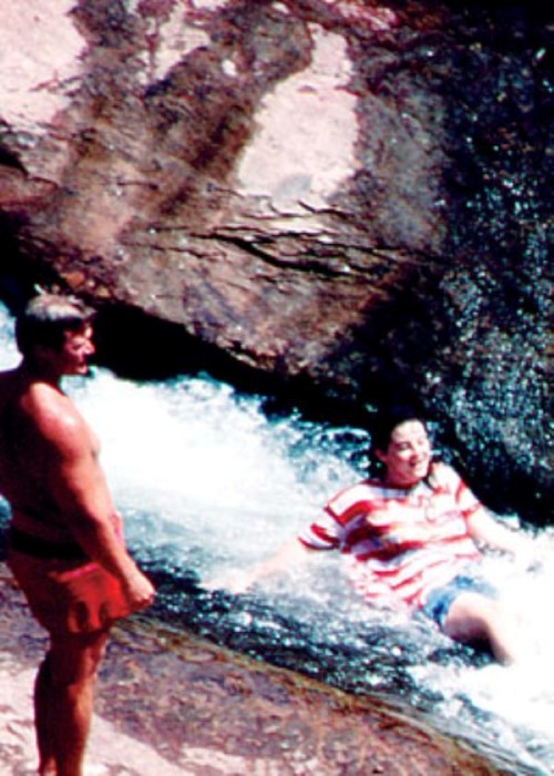 Two people near a rocky stream; one is standing while the other is sitting in the water, enjoying the moment.