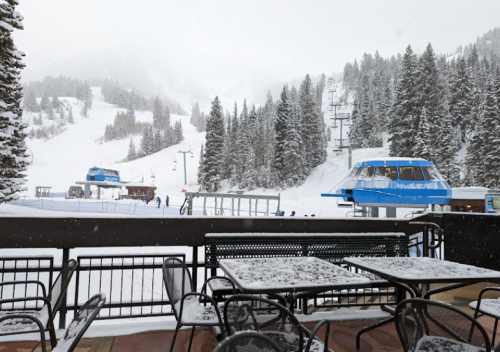 Snow-covered ski resort with blue gondolas, trees, and empty tables in the foreground. Snowflakes are falling.
