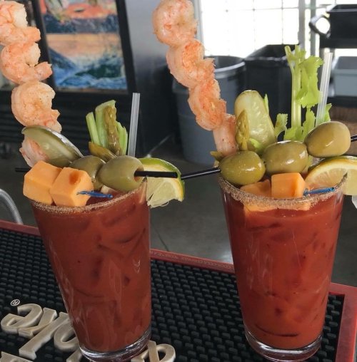 Two colorful Bloody Mary cocktails garnished with shrimp, celery, olives, lime, and cheese cubes on a bar counter.