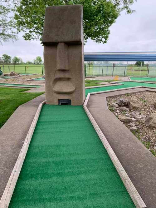 A mini-golf course featuring a large stone Moai statue at the end of a green carpeted path.