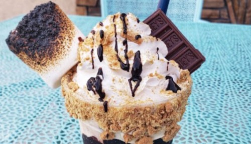 A decadent dessert featuring whipped cream, chocolate drizzle, a toasted marshmallow, and graham cracker crumbs.
