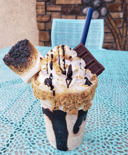 A decadent milkshake topped with whipped cream, chocolate drizzle, a toasted marshmallow, and a chocolate bar on a blue table.