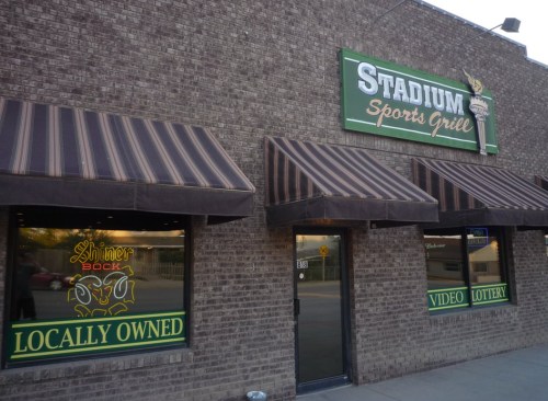 Exterior of Stadium Sports Grill with striped awnings, signs for video lottery, and "Locally Owned" displayed.