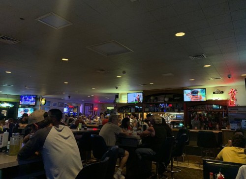 A busy bar with patrons seated at the counter, watching TVs and enjoying drinks in a dimly lit atmosphere.