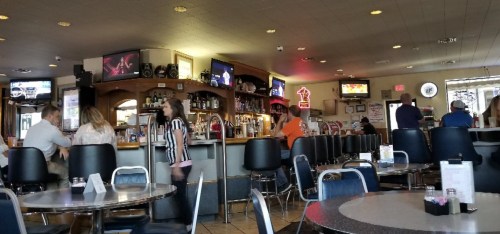 A casual diner interior with a bar, patrons seated at tables, and multiple TVs showing sports.