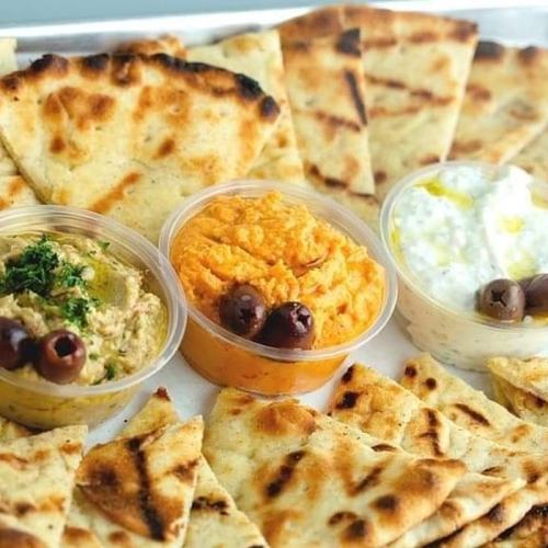 A platter of pita bread with three dips: hummus, a spicy orange spread, and a yogurt dip, garnished with olives.