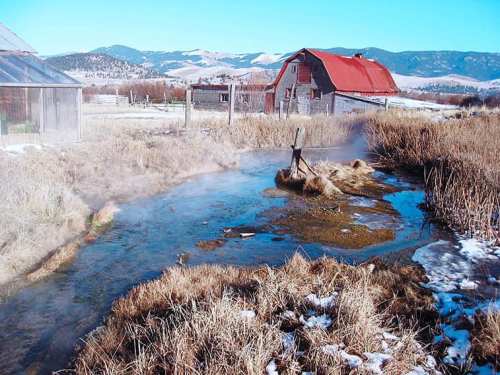A serene landscape featuring a steaming stream, dry grass, and a rustic barn with a red roof against a mountain backdrop.