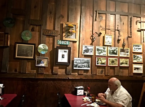 A rustic restaurant interior with wooden walls, framed photos, and a man eating at a table. Sunflowers decorate the space.