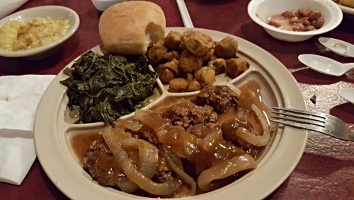 A plate of Southern comfort food featuring meat with onions, collard greens, fried okra, and a roll.