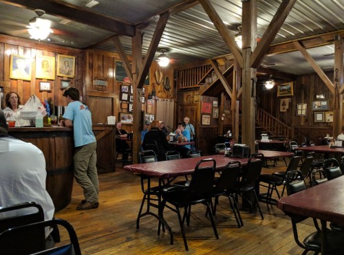 A rustic restaurant interior with wooden walls, tables, and patrons dining. A bar area is visible in the foreground.