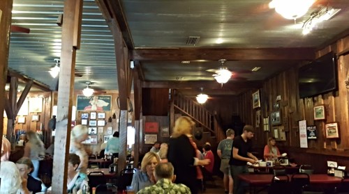 A bustling restaurant interior with wooden walls, tables, and patrons enjoying their meals. Fans and decor visible.