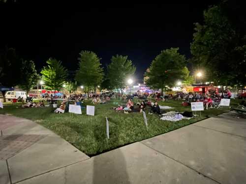 A crowd gathers on a grassy area at night, surrounded by trees and food trucks, enjoying an outdoor event.