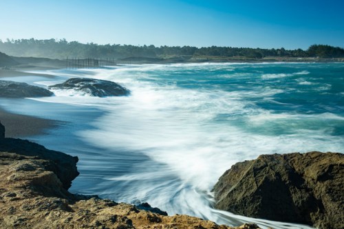 Waves crash against rocky shore under a clear blue sky, with distant trees and a beach in the background.