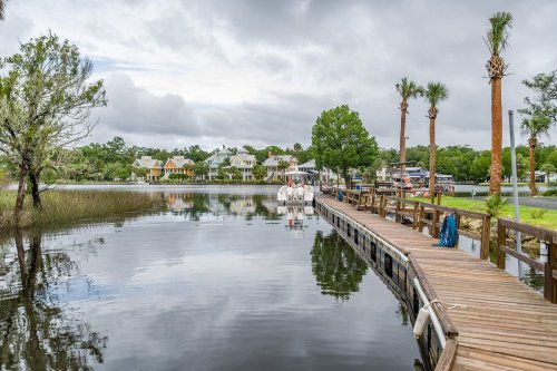 A wooden dock extends into a calm lake, surrounded by lush greenery and colorful houses under a cloudy sky.