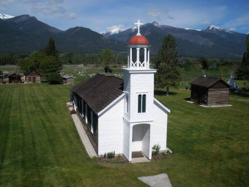 A white church with a red dome, surrounded by green grass and mountains under a blue sky.