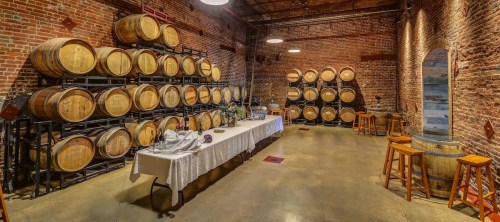 A rustic wine cellar with stacked barrels, a long table set for dining, and wooden stools against a brick wall.