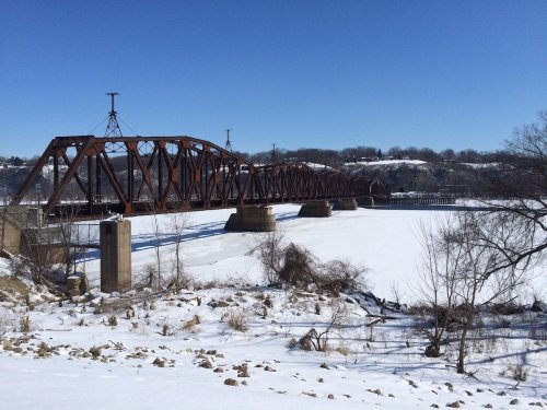 A rusty train bridge spans a frozen river, surrounded by snow-covered land and trees under a clear blue sky.