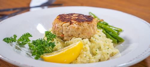 A plate featuring a crab cake on creamy risotto, garnished with lemon, green beans, and parsley.