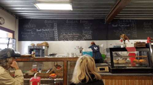 A café menu board displays sandwich options and prices, with customers seated in front. A staff member is preparing food.