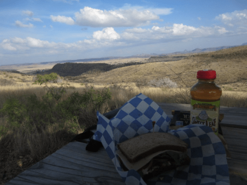 A sandwich and a bottle of tea on a picnic table, overlooking a vast, scenic landscape with clouds in the sky.