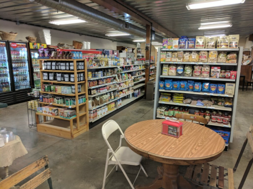 Interior of a grocery store with shelves stocked with various food items and a small table with chairs.