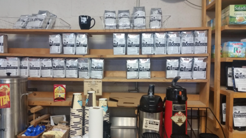 A wooden shelf displaying various bags of coffee, cups, and coffee equipment in a cozy shop setting.