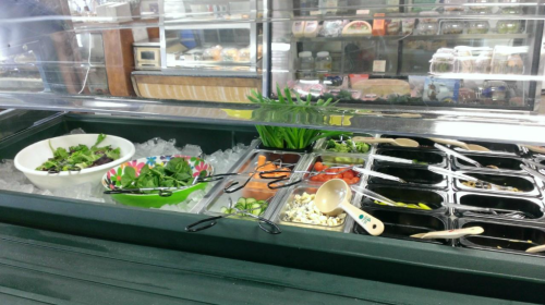 A salad bar with fresh greens, tomatoes, and toppings displayed on ice in a refrigerated case.
