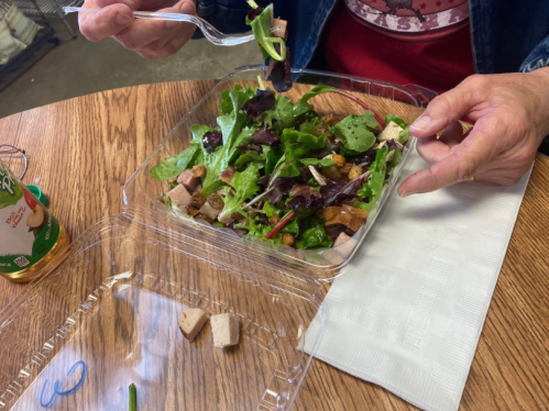 A person holds a fork over a salad in a clear container on a wooden table, with a drink and napkin nearby.