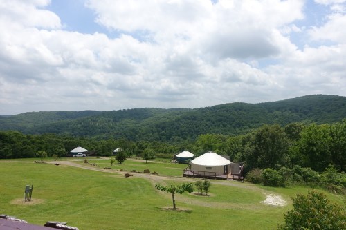A scenic view of green hills with several yurts and a wooden deck under a partly cloudy sky.