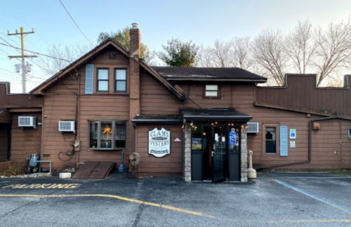 A rustic brown building with a sign reading "Clams & Oyster," featuring a parking area and trees in the background.