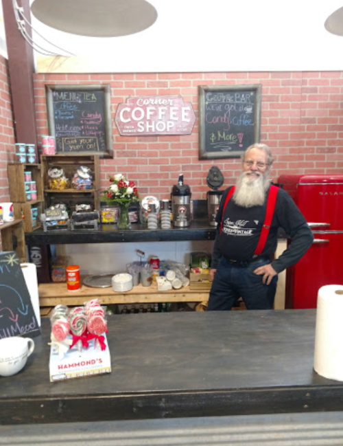 A bearded man in a red shirt and suspenders stands behind a coffee shop counter with snacks and decorations.