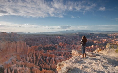 A person stands on a rocky ledge, overlooking the stunning orange rock formations of Bryce Canyon under a blue sky.