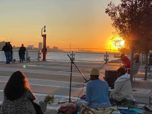 People enjoying a sunset by the water, with musicians performing in the foreground and a city skyline in the background.