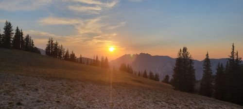 Sunset over a mountainous landscape, with silhouettes of trees in the foreground and a warm, colorful sky.