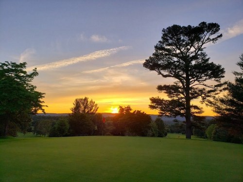 A serene golf course at sunset, with vibrant colors in the sky and a silhouette of trees in the foreground.