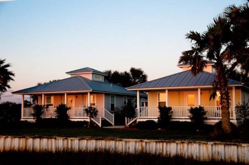 Two beach houses with metal roofs, surrounded by palm trees, at sunset with a calm atmosphere.