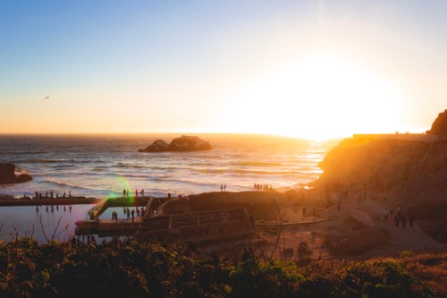 Sunset over the ocean with silhouettes of people along the shore and rocky formations in the background.
