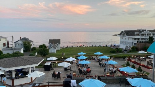 A scenic view of a beach at sunset, with umbrellas and diners in the foreground and people along the shoreline.