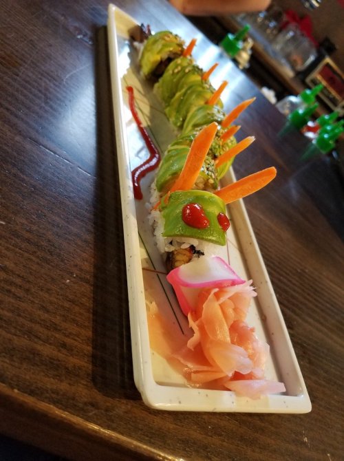 A colorful sushi roll shaped like a caterpillar, topped with avocado, carrots, and served with pickled ginger.