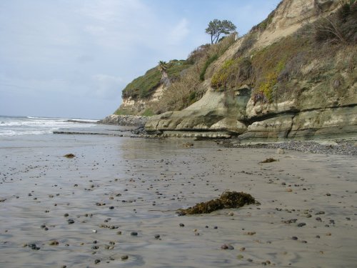 A serene beach scene with rocky cliffs, gentle waves, and scattered pebbles along the shore under a cloudy sky.