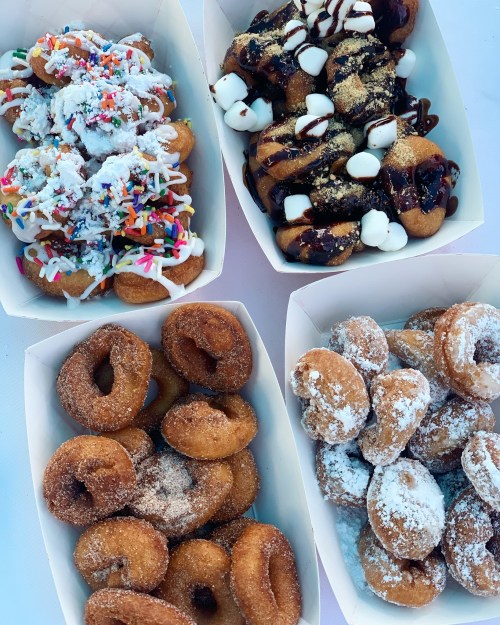 Four trays of assorted donuts: some with sprinkles, chocolate drizzle, and powdered sugar, showcasing a variety of flavors.