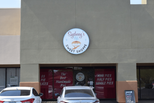 Exterior of Sydney's Sweet Shoppe, featuring a sign and red curtains, with parked cars in front.