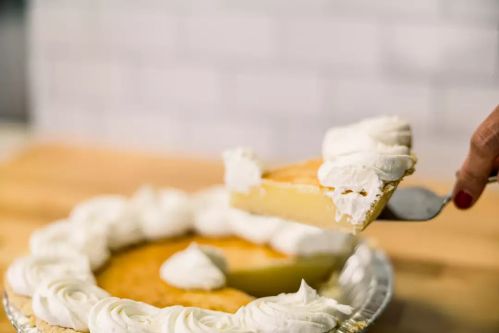 A slice of pie being lifted from a whole pie topped with whipped cream on a wooden surface.
