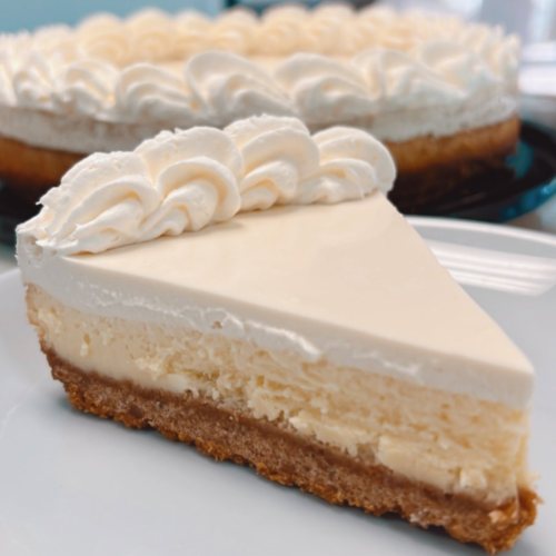 A slice of creamy cheesecake with a graham cracker crust and whipped cream topping, served on a white plate.