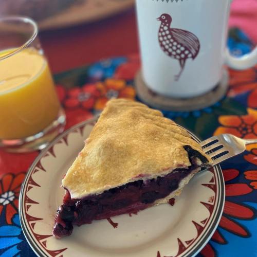 A slice of berry pie on a decorative plate, with a glass of orange juice and a mug in the background.