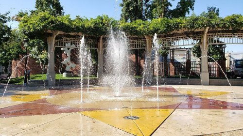 A fountain sprays water in a sunny park, surrounded by greenery and a decorative structure.